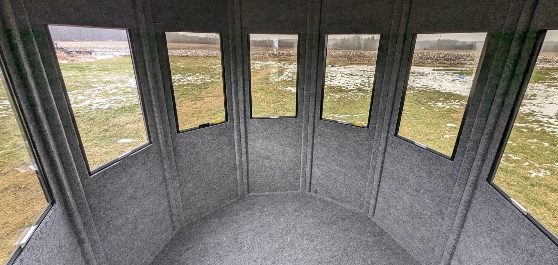 Inside a NXTGen 6 Foot blind: carpeted walls, tinted vertical-slide windows in every panel, field beyond.